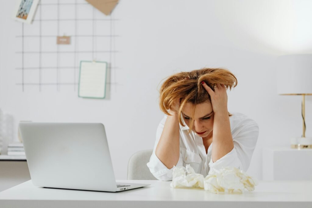 Frustrated woman in white shirt at desk with laptop, feeling overwhelmed.